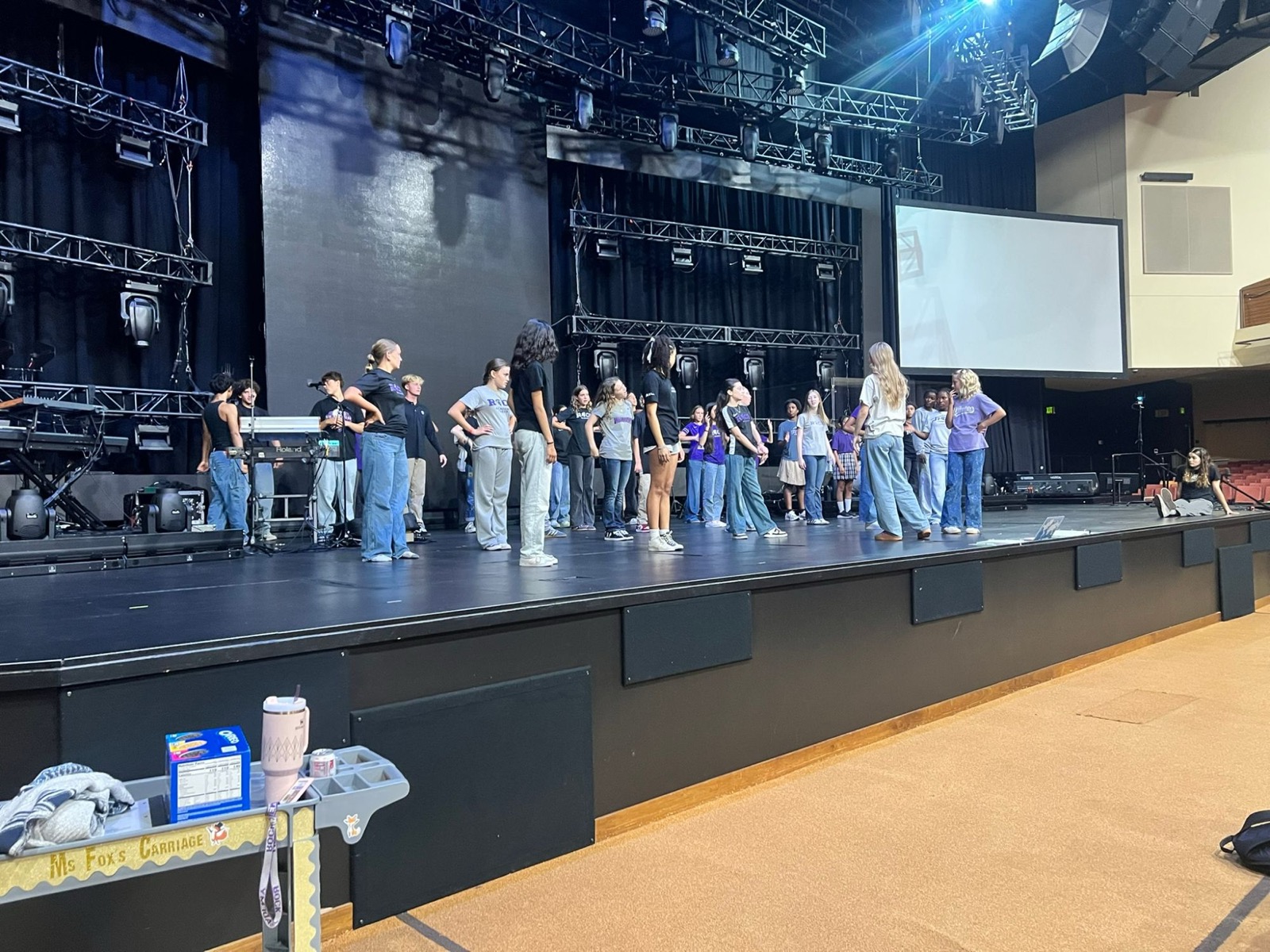 Students in rehearsal on a large stage, with a supply cart labeled Ms. Fox's Carriage in the foreground