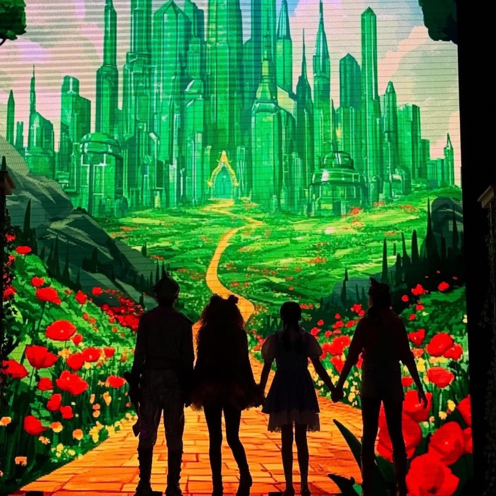 Four Wizard of Oz leads silhouetted in front of the Emerald City, holding hands on the yellow brick road